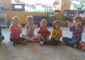 Biblioterapia w grupie 5. Grupa dzieci siedzi w kółku, pokazując gest podniesionych rąk na znak wspólnego działania.